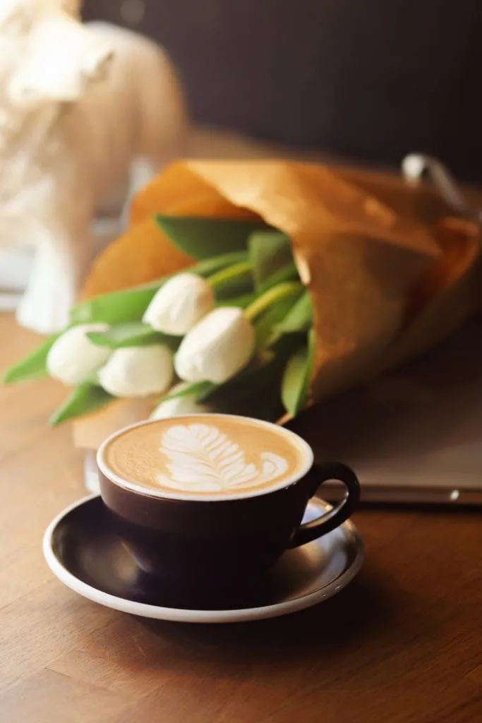 Cappuccino mit Latte Art in dunkler Tasse, dahinter ein Strauß weißer Tulpen