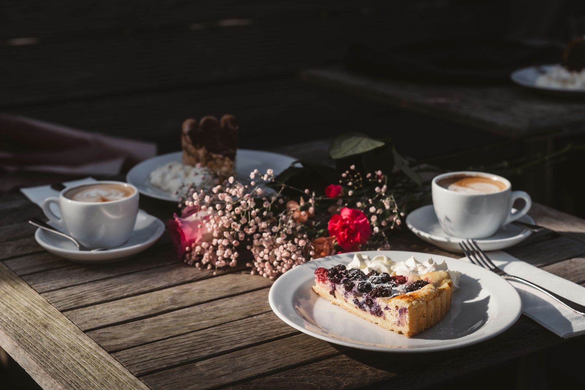 Zwei Cappuccino, Blumen und ein Stück Kuchen auf einem Holztisch im Garten